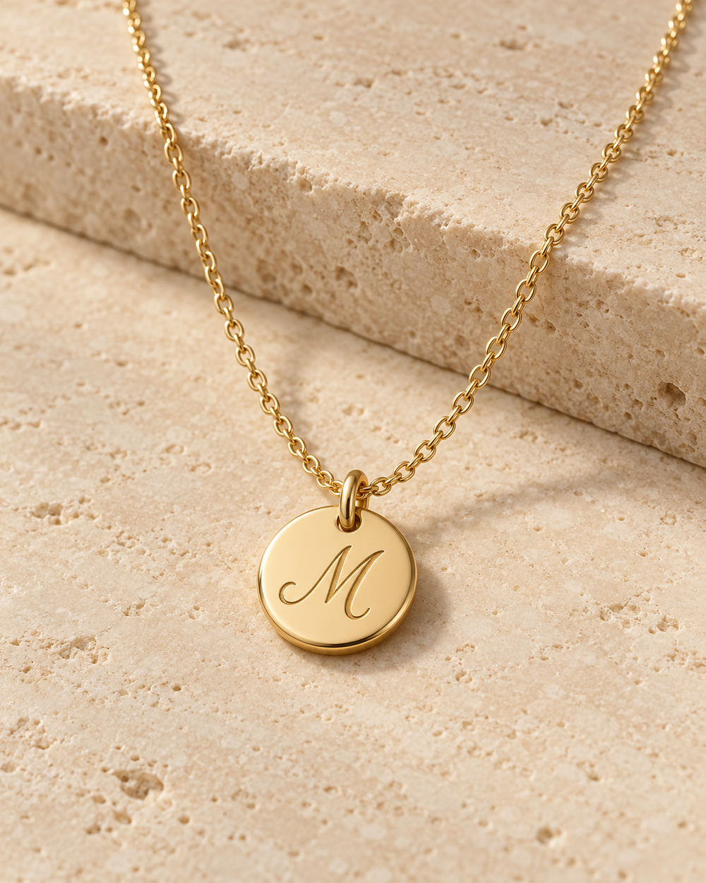 Illustrative gold M initial pendant concept with gold chain on cream stone